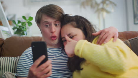 Una-Pareja-Joven-Con-Síndrome-De-Down-Sentada-En-Un-Sofá-Usando-Un-Teléfono-Móvil-Para-Tomarse-Un-Selfie-En-Casa