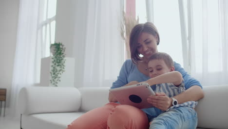 Madre-E-Hijo-Sentados-En-Un-Sofá-Usando-Una-Tableta-Digital.-Mamá-Feliz-Y-Niño-Pequeño-Usando-Una-Tableta-Con-Pantalla-Táctil-Juntos-Viendo-Un-Video.-Madre-Sonriente-Y-Niño-Lindo-Jugando-En-Una-Tableta-Digital.