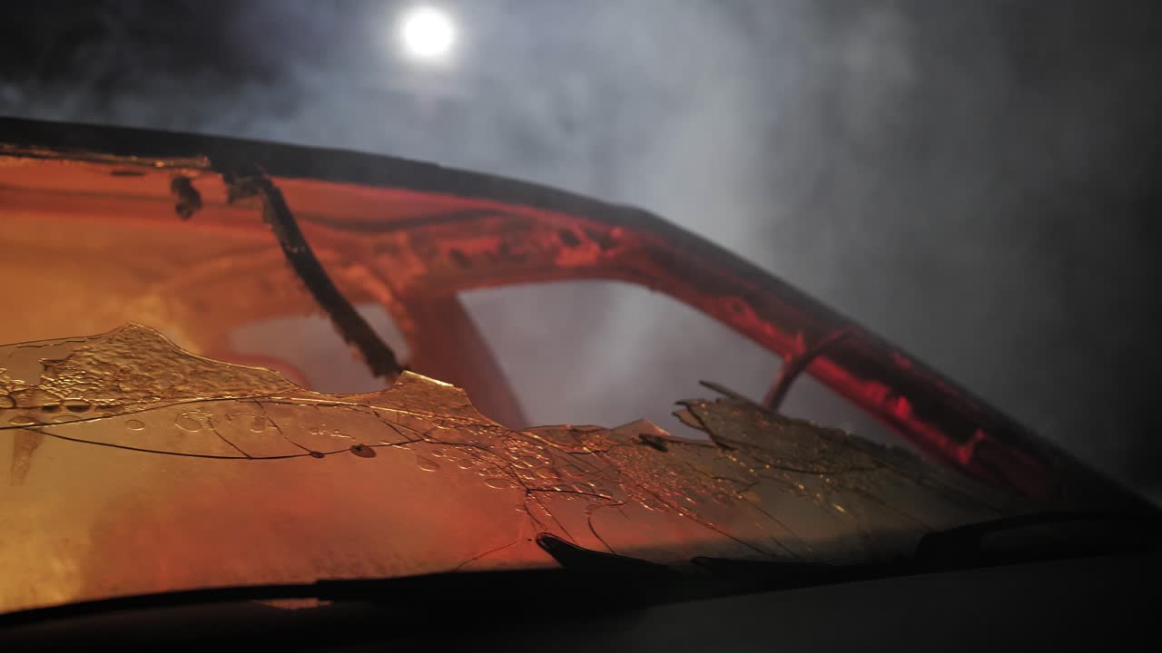 Slow motion footage of a shattered windshield in a burnt car, captured under eerie night lighting with smoke