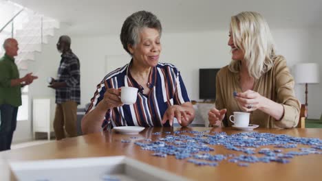 mujeres mayores afroamericanas y caucásicas sentadas a la mesa haciendo rompecabezas bebiendo té