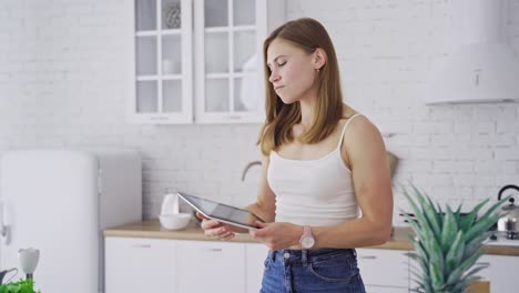 Beautiful woman on a white kitchen. Young model is looking into recipe on the tablet to prepare healthy food at home.