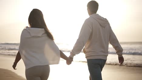 backside view of young couple in love holding hands walking on the beach in casualclothes. background sunset in sea. slow motion. mild dusk