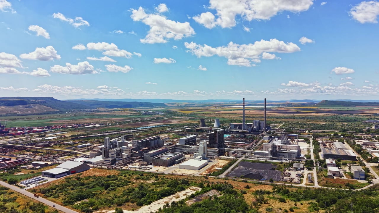 Vista aerea di un complesso industriale con fabbriche, centrali elettriche e campi in una valle, sotto un cielo azzurro e limpido con nuvole durante il giorno.