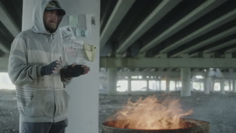 homeless man warming hands by fire barrel