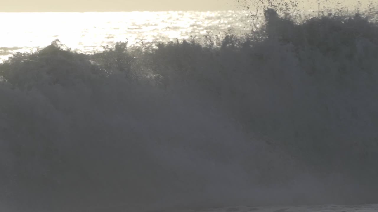 Powerful ocean waves crashing near the shore of Puerto Escondido, bathed in the bright, hazy light of a sunset