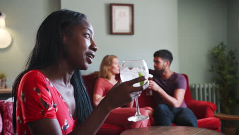 Close-up-of-young-black-woman-sitting-in-the-lounge-at-a-pub,-holding-a-drink-and-talking-to-her-friend,-side-view