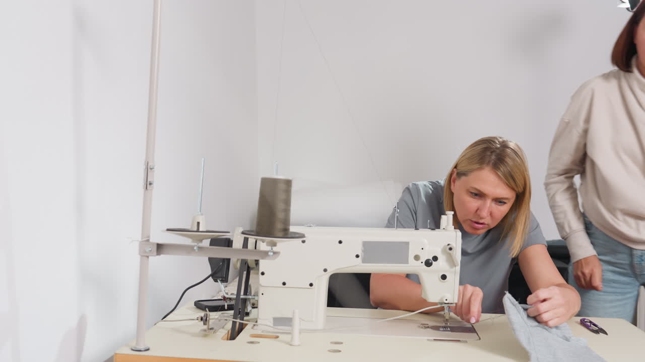 Fashion designers adjusting needle on bar tack sewing machine, one helps partner then returns to seat, showing synergy, professional environment, shared skill, efficient production