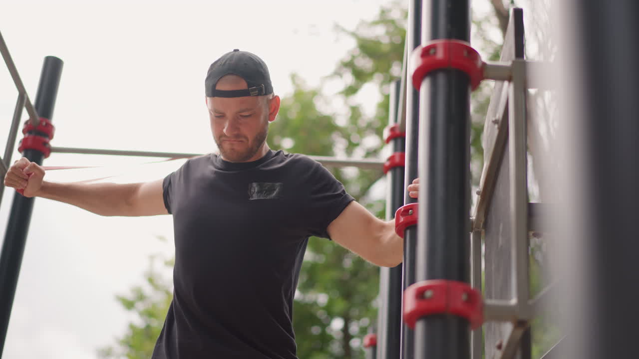 Man Warming Up With Resistance Band At Outdoor Calisthenics Park, Black Shirt And Backward Cap, Focused Expression While Pulling Band Across Chest, Metal Bars With Red Connectors, Preparing