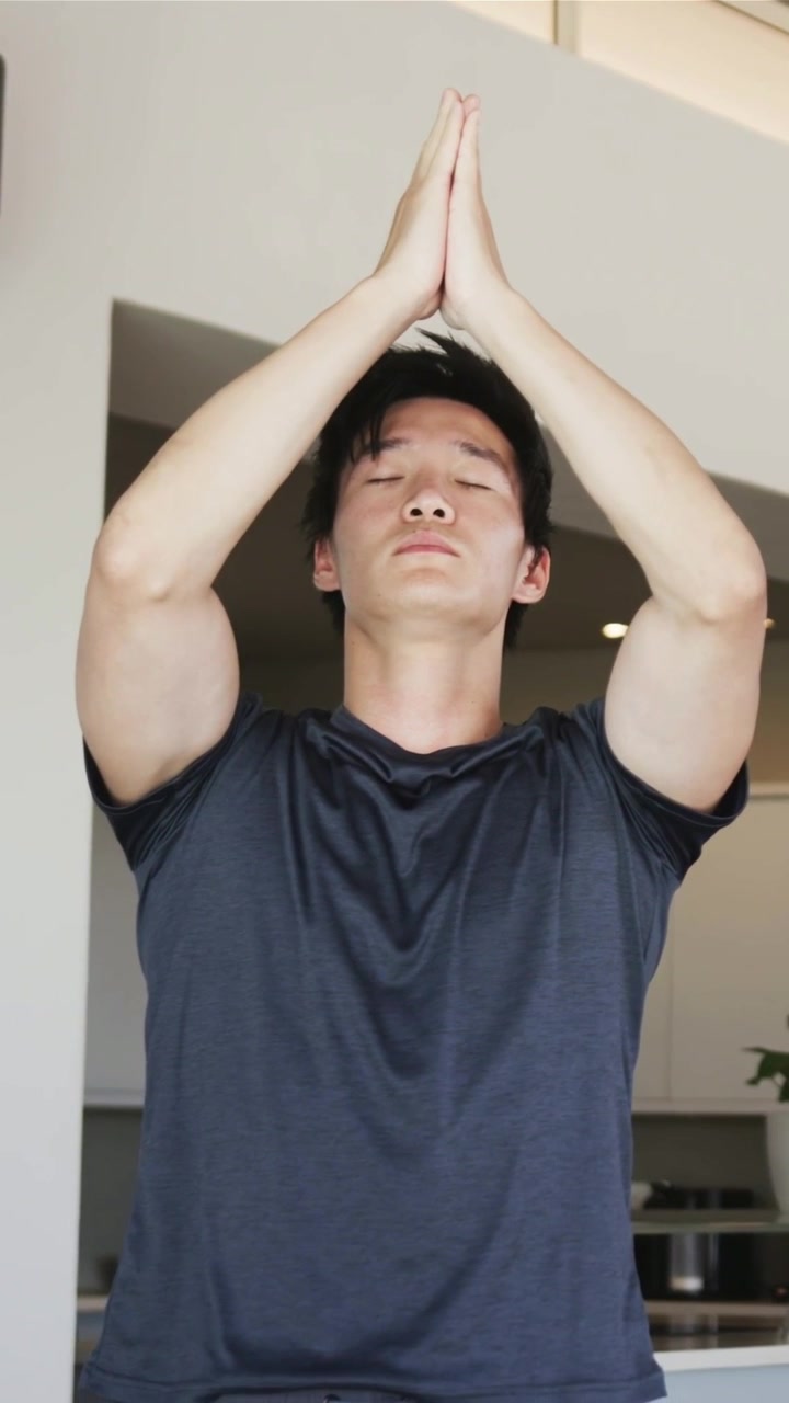 Vídeo vertical: Homem de camiseta escura abaixando-se, trazendo as mãos de cima da cabeça para o peito na cozinha para ioga.