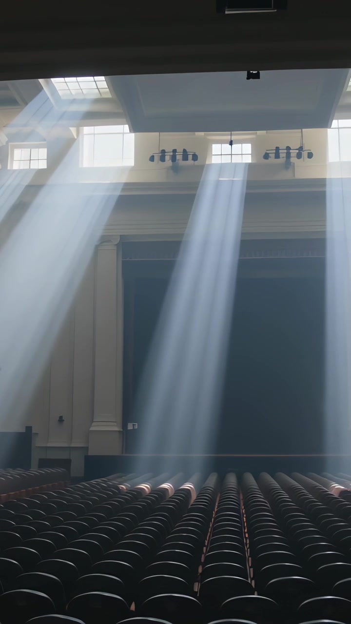 Video verticale: Raggi di sole che filtrano e illuminano file di sedie scure in un grande auditorium, provenienti da lucernari.