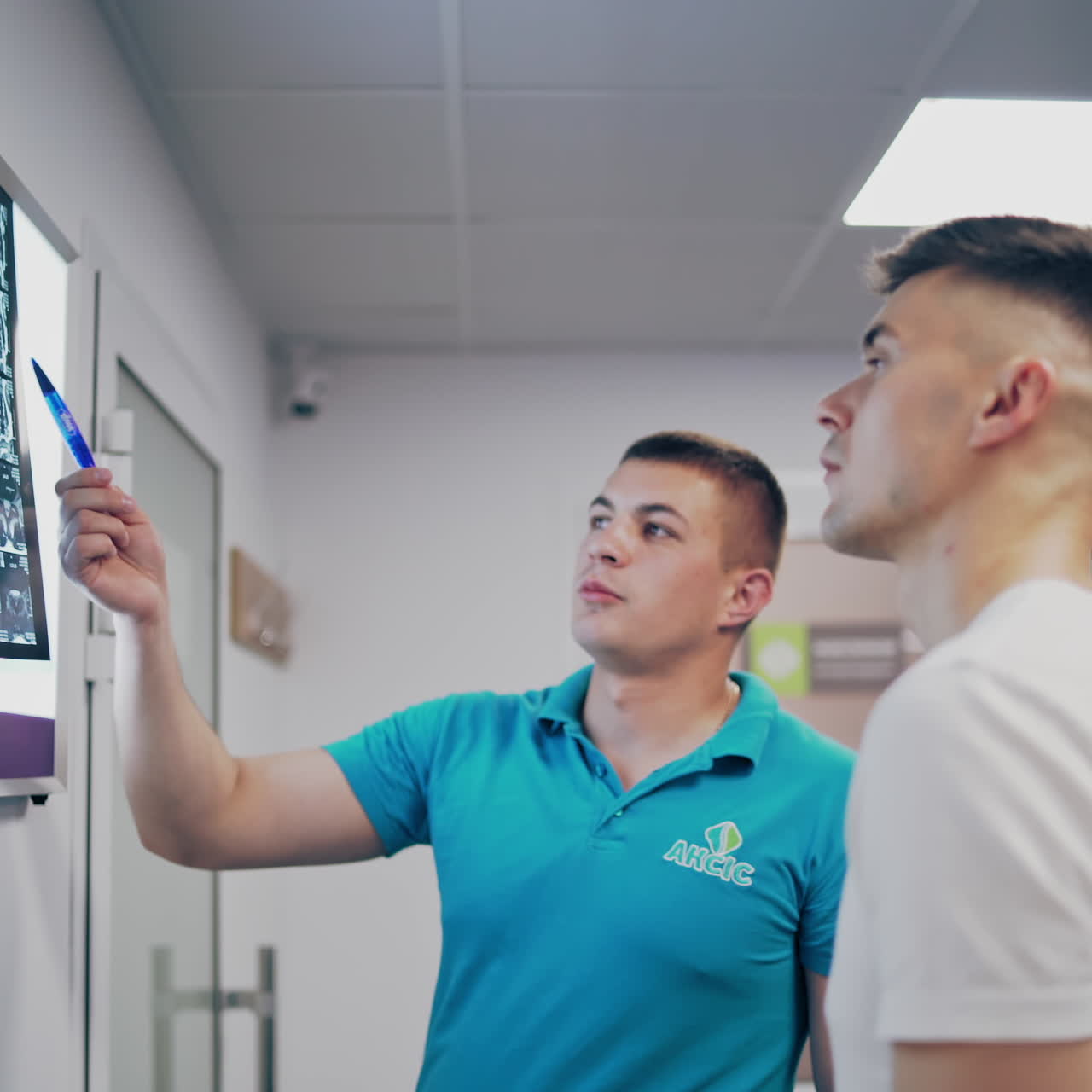 Young doctor shows the problem areas of the back on the X-ray to the patient. Therapist man talks to a patient and shows spine x-ray on a white board in clinic.