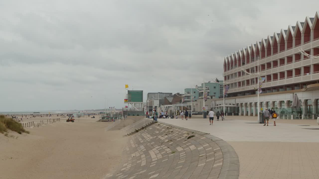 People walk on seaside promenade beside sandy beach, modern buildings, overcast sky, wide static shot