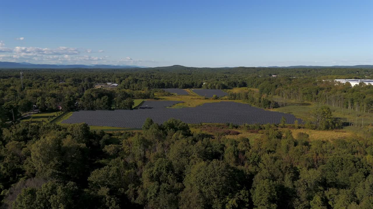 A high angle, aerial view over a large field with trees on a sunny day with clear blue skies. The drone camera slowly dolly in and boom up over the trees to reveal many solar panels