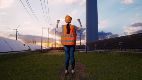 full body back view of a female engineer with safety helmet raising her hands celebrating while working with solar panel and wind turbines