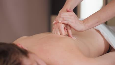 massage therapist folding hands to apply focused pressure on back of relaxed client lying on white sheet-covered massage table, close-up detail showing hand movement and therapeutic massage technique