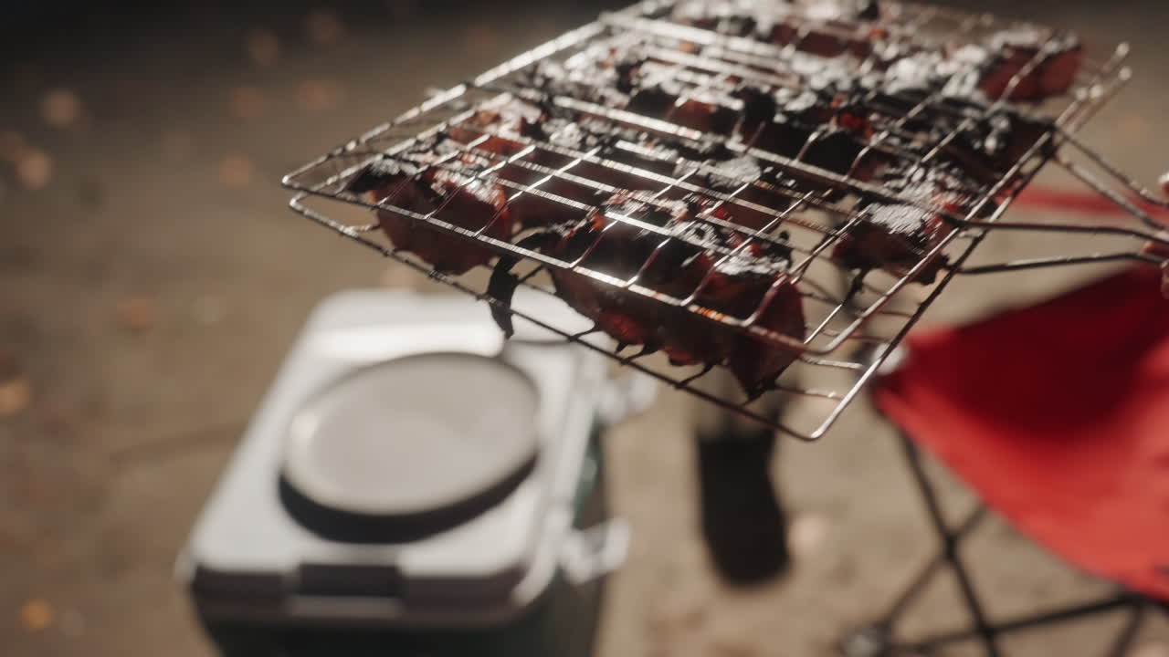Primo piano di spiedini che grigliano sopra un frigo portatile, con una griglia appoggiata su un frigo di polistirolo, accanto a una sedia pieghevole rossa. Luce calda serale, carne caramellata con bordi bruciacchiati, fumo leggero e sfrigolio, cuoco solitario rilassato.