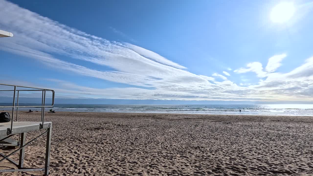 Camera pans from lifeguard hut to wide sandy beach under bright sunlight and dramatic clouds