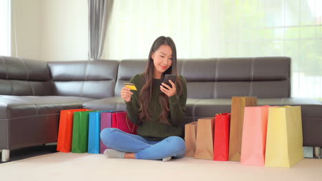 a pretty, young woman sitting on the floor in front of a couch surrounded by colorful shopping bags checks and rechecks her credit card number on a smartphone