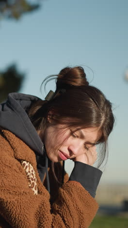 una chica se sienta sola en un banco en un parque urbano, con el corazón pesado y la mano apoyada en la cara. está vestida con un abrigo marrón, pantalones vaqueros azules y zapatos blancos, con una mochila negra colocada al lado