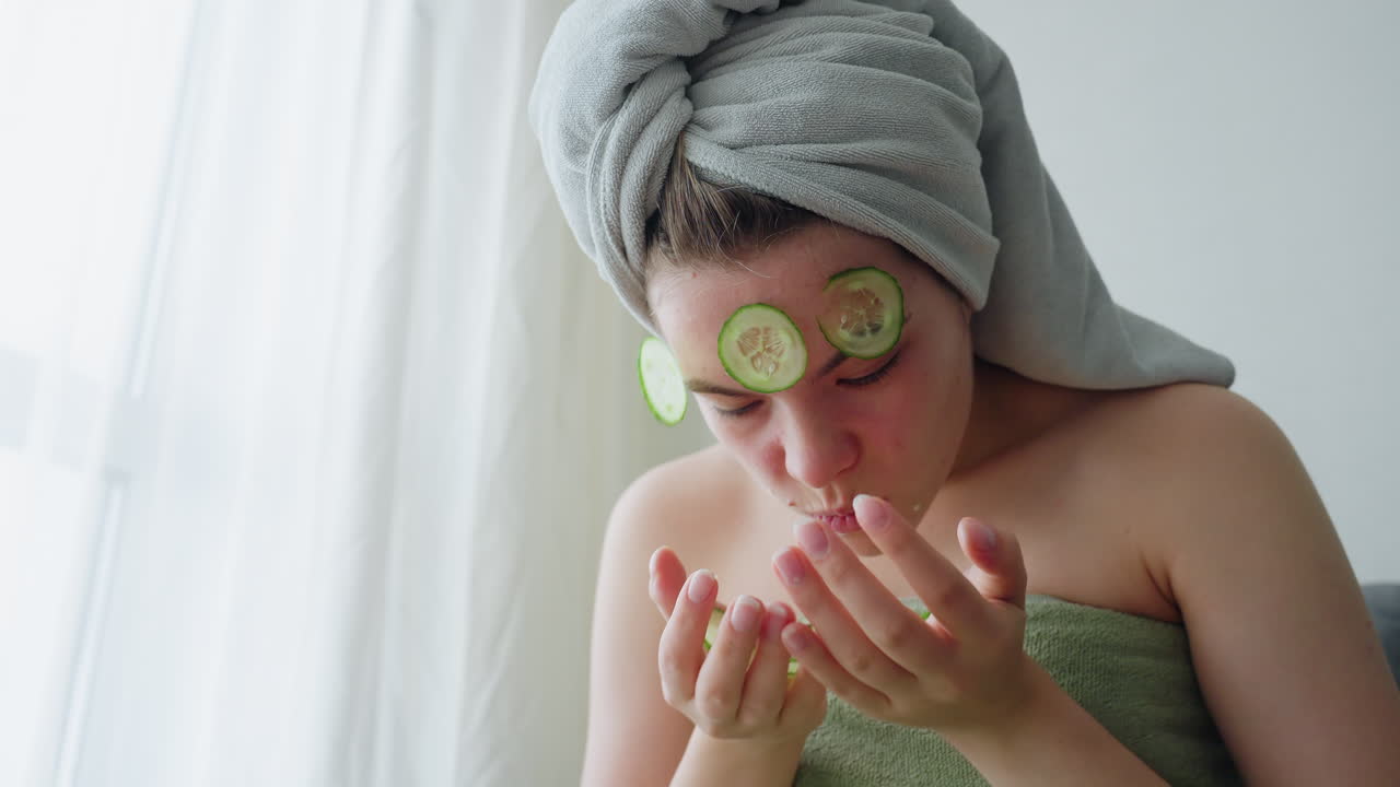 Close view of girl wrapped in towel squeezing face playfully while removing cucumber slices from skin during refreshing skincare moment, soft natural light enhances calm, peaceful spa-like atmosphere