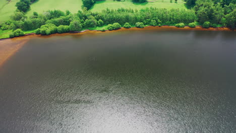 Aerial-shot-of-a-lake-in-the-bright-sunshine-with-the-water-glistening,-lake-edge-has-a-green-forest
