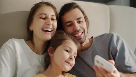 A-happy-brunette-man-in-a-gray-T-shirt-and-his-cheerful-wife,-a-brunette-girl-in-a-white-T-shirt,-together-with-their-daughter,-a-little-girl-in-a-yellow-dress,-take-a-selfie-and-look-at-the-screen-of-a-White-phone-while-sitting-on-a-large-light-brown-sofa