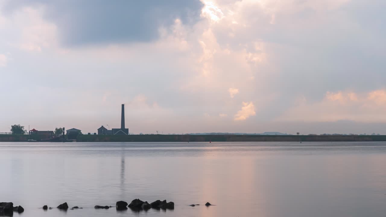Silhouette of an old steam pumping station across calm water at sunset with dramatic clouds and pastel reflections. Timelapse.