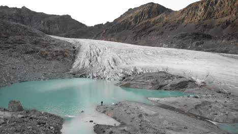 Aerialf-flyover-over-a-hiker-at-the-shore-of-the-turquoise-glacial-lake-at-Hohlaub-glacier-near-Saas-Fee-in-Valais,-Switzerland