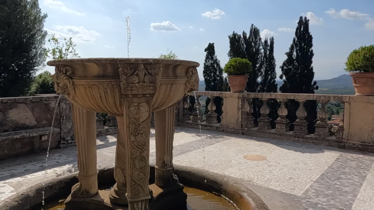 Roma, İtalya yakınlarındaki tarihi Tivoli villasında, güneşli bir terasta nazikçe akan zarif oyma taş çeşme. Arka planda klasik mimari, selvi ağaçları ve yuvarlanan tepeler.