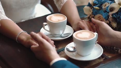 newlyweds hold hands while sitting in a cafe sitting at a table on which there are two mugs of coffee close-up