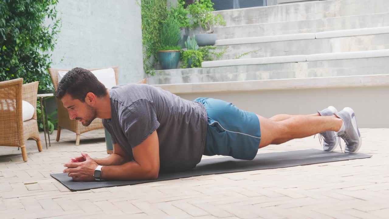 Man placing forearms, holding plank, tightening core for strength on patio in shorts with mat
