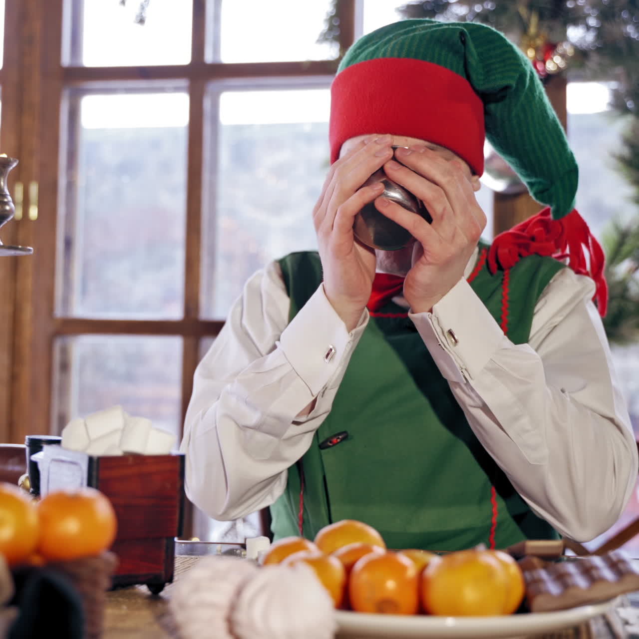 Funny elf with drinks and becomes dirty on his face on Christmas background. Hungry male elf in red and green costume drinks at festive table indoors. Close-up.