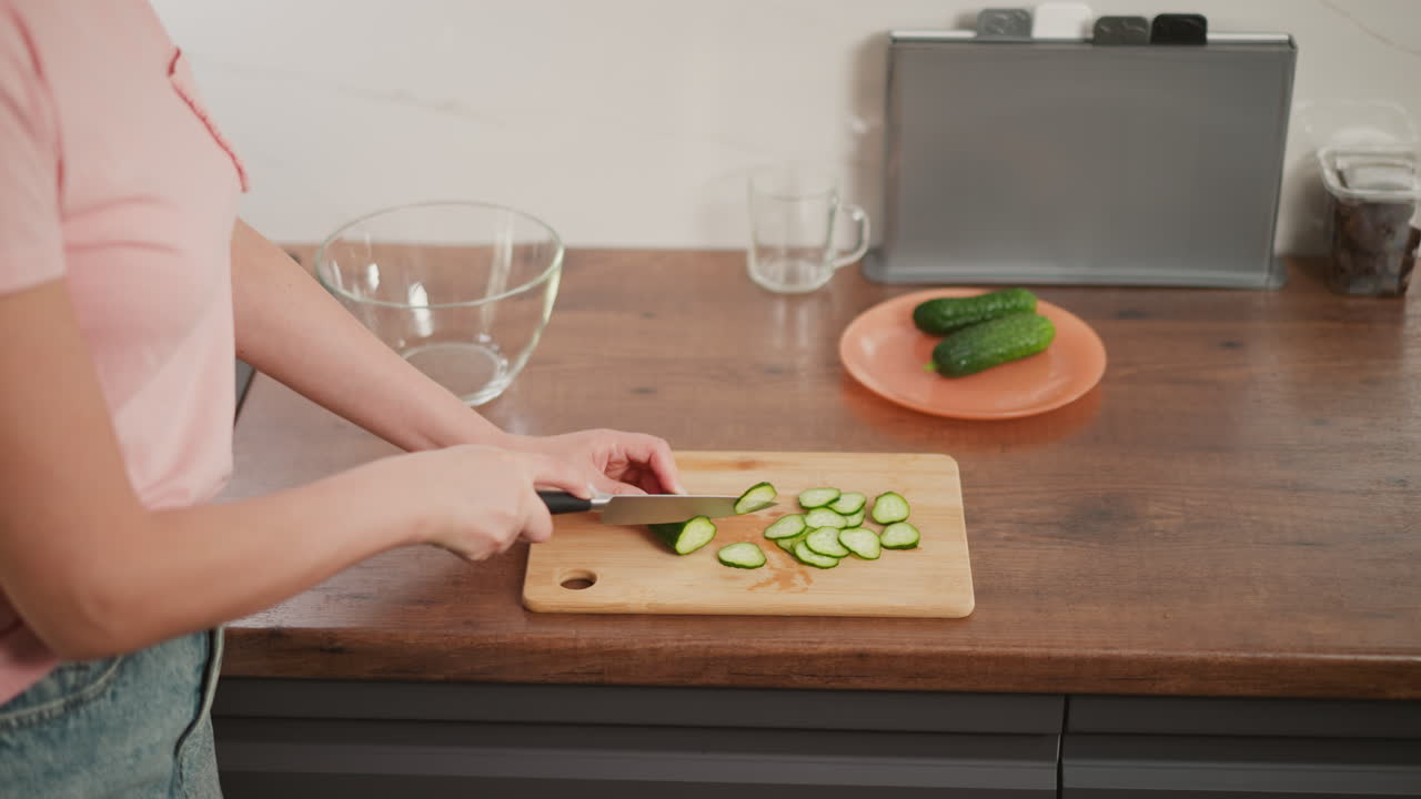 Woman slicing cucumber into thin pieces on wooden cutting board with two whole cucumbers on orange plate in kitchen setting, clear glass bowl and utensils placed on clean wooden counter