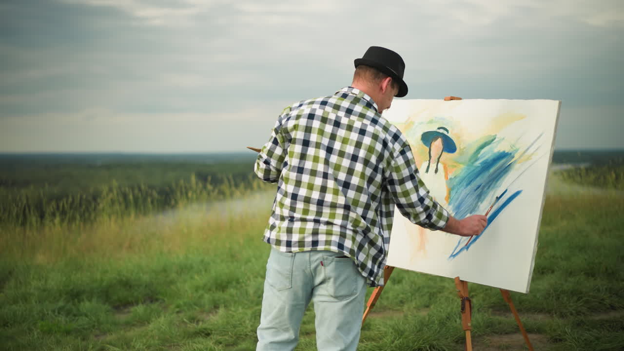 Back view of an artist wearing a black hat and checkered shirt, holding a palette while painting a large, blue-themed canvas in a peaceful grass field