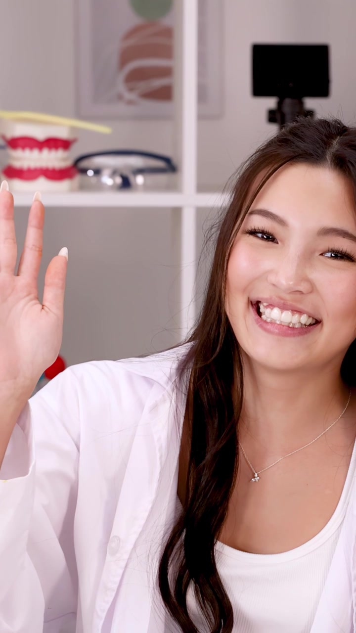 A happy student in a white lab coat waves at the camera in a classroom.