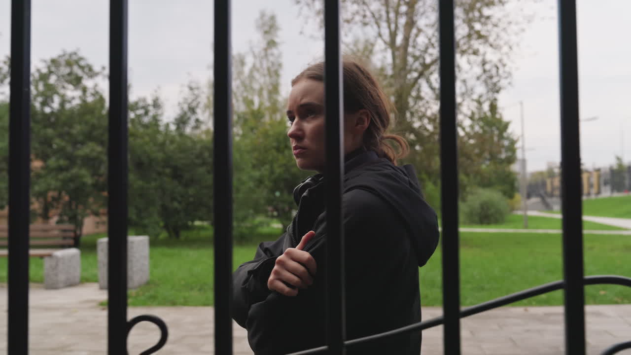 Woman behind iron fence in park, slow walk with arms folded, distant gaze toward trees, overcast sky and damp pavement, vertical bars framing profile, candid cinematic portrait, introspective mood