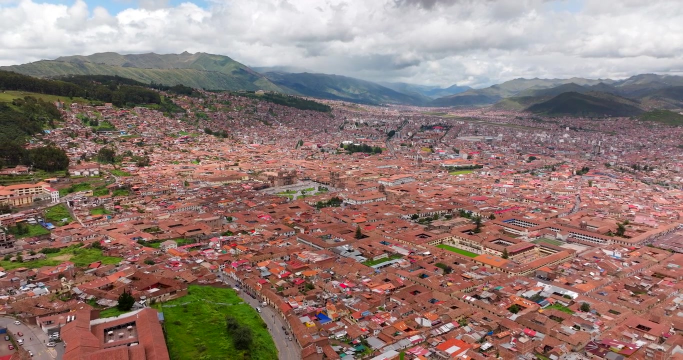 Drone aerial view over Cusco, Peru, showing dense urban architecture surrounded by Andean mountains under dramatic cloud formations in South America