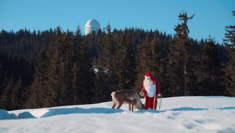 Santa-Claus-Y-Uno-De-Sus-Ciervos-Están-Parados-En-Una-Ladera-Nevada