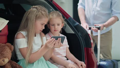 two caucasian children browsing mobile phone while sitting in car trunk.