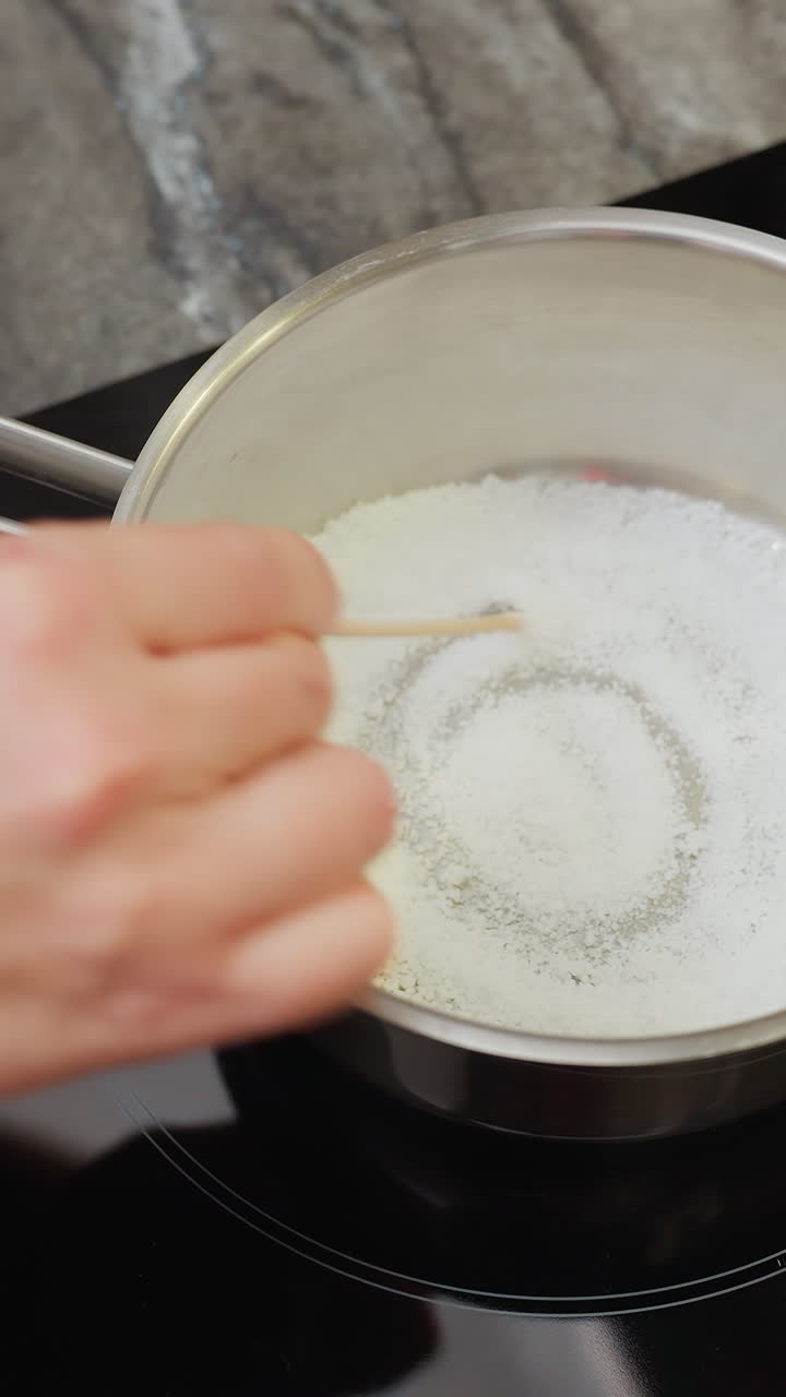 primer plano de una persona mezclando azúcar en una olla con una cuchara de madera, acción de cocción en la cocina, preparación de ingredientes para caramelo o postre en la estufa, azúcar en la sartén calentándose