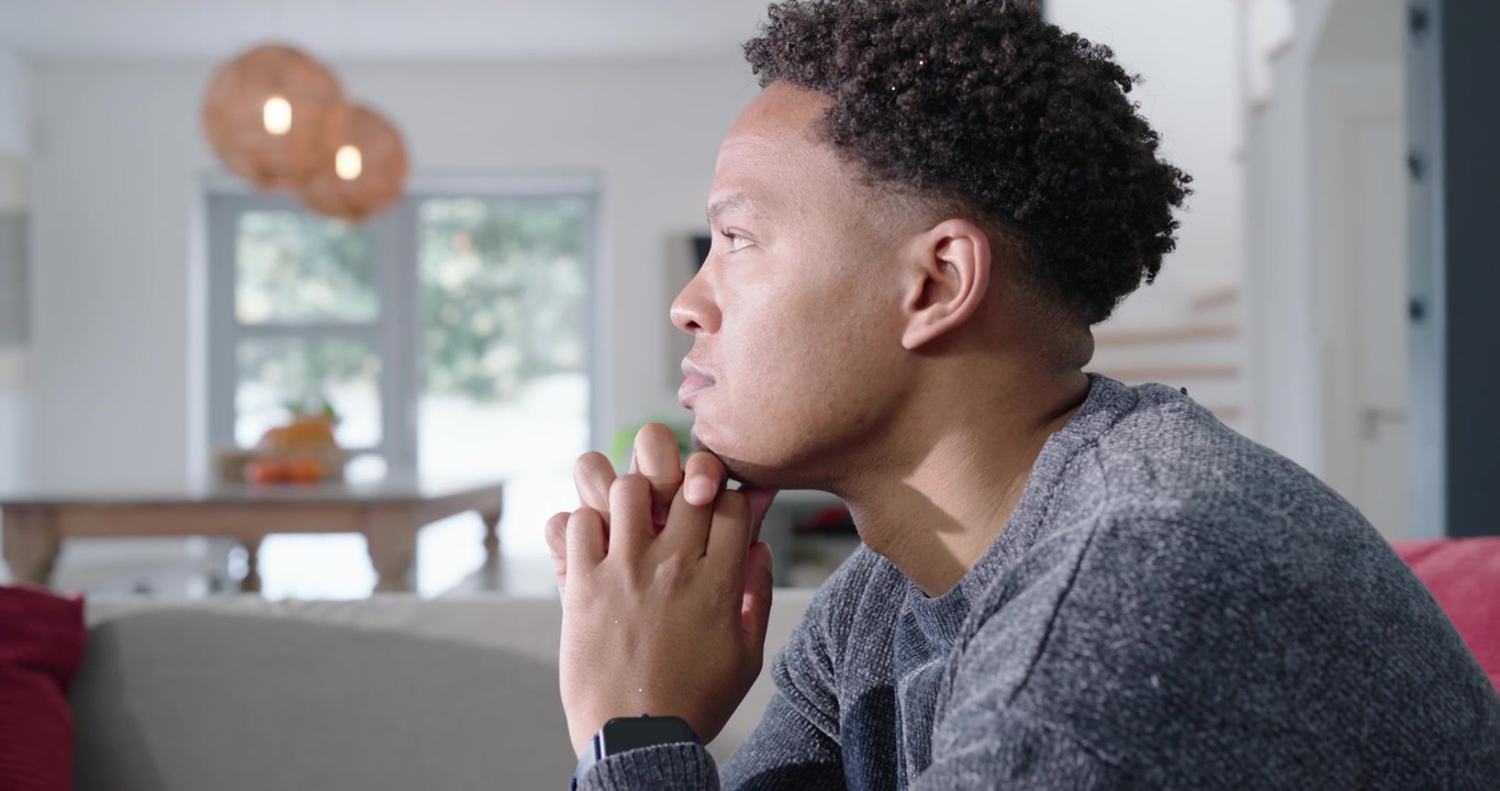Câmera recuando, homem afro-americano adulto no sofá, apoiado nas mãos, em reflexão, com espaço para texto.