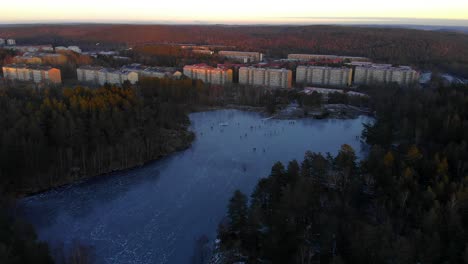 Toma-Aérea-De-Personas-Patinando-Sobre-Hielo-En-Un-Lago-Congelado,-Hermoso-Pueblo-En-Una-Escena-De-Invierno-Al-Amanecer