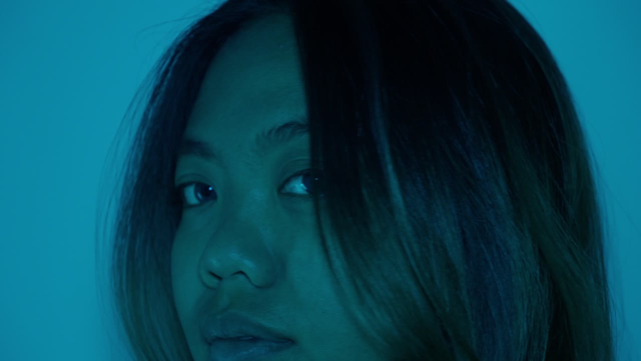 Woman in blue lighting with contemplative expression, looking downward in quiet reflection turns to face forward slowly