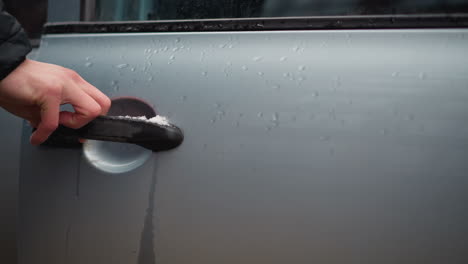 close up motorist opening door handle on ash coloured car with metallic finish and water droplets and snow on panel surface during winter on urban street lined with parked vehicles with reflection
