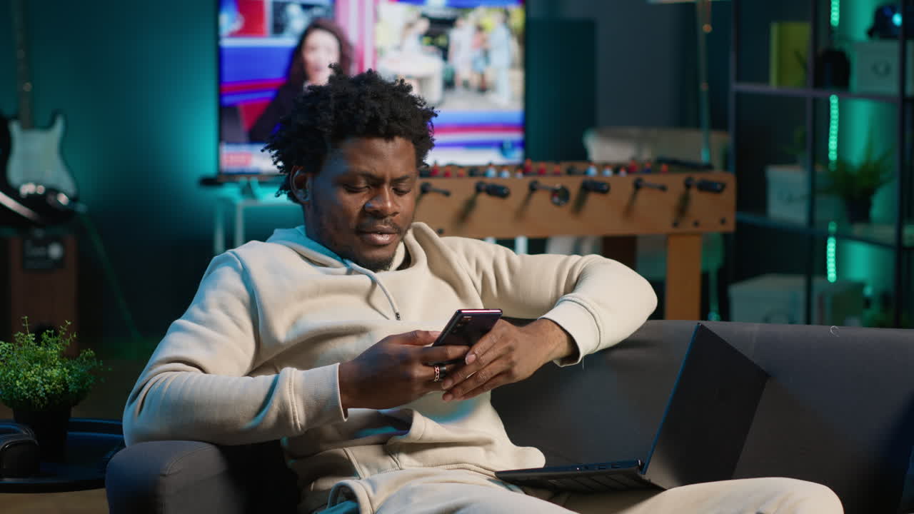 Relaxed African American freelancer smiling while browsing his smartphone on a comfortable couch. Modern home setting highlights leisure and online connectivity in a stylish apartment. Camera A.