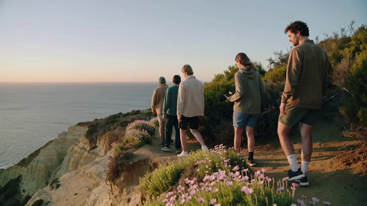 Walking five hooded friends along bluff at dusk to view ocean front pointing middle filming phone