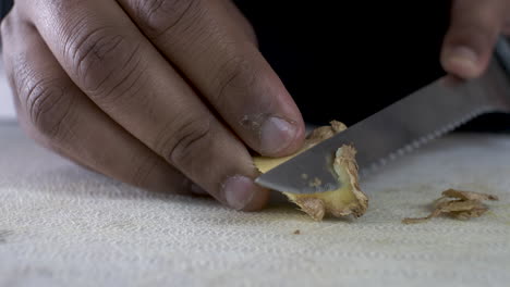 hand of ethnic minority adult male using serrated knife to peel skin off ginger on cutting board