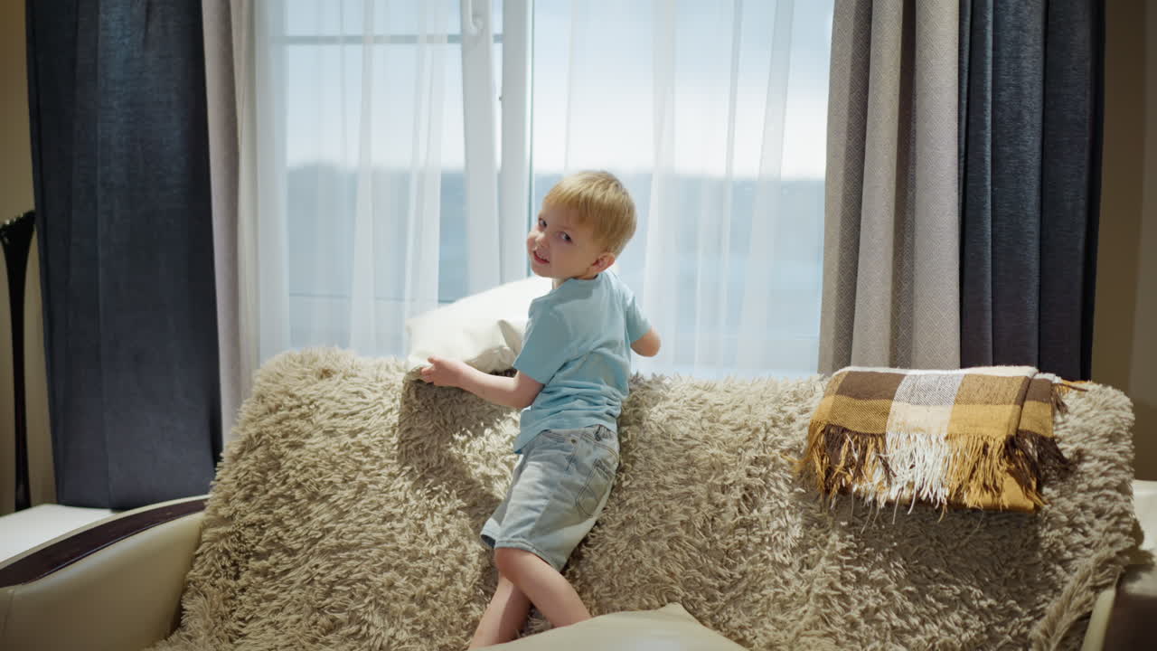 Smiling young boy in blue shirt sitting on soft sofa holding cushion, playful cheerful expression, cozy living room interior, warm blanket and throw in background, carefree childhood moment of joy, laughter
