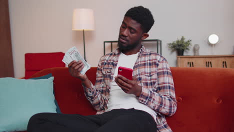 Sad-poor-african-american-man-holding-cash-money-and-mobile-phone-calculate-domestic-bills-at-home