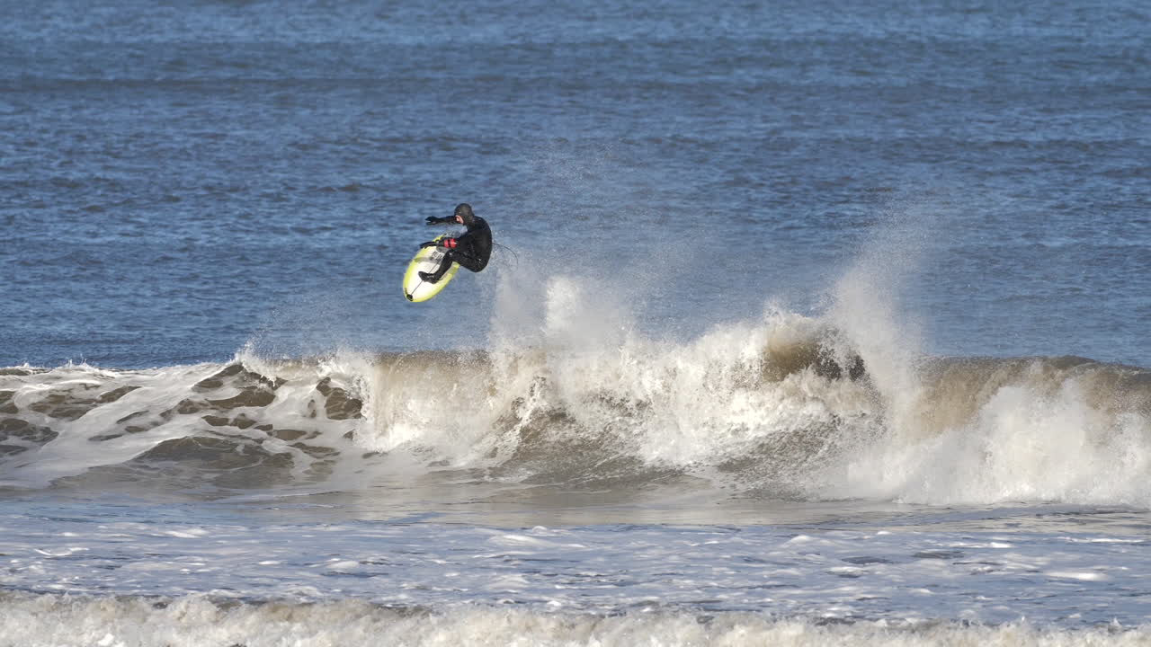 Male advanced level surfer in black hooded wetsuit on a mid-height wave and completing an aerial manoeuvre, in cold water conditions on a bright and sunny day.